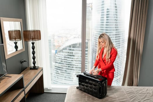 Young blond businesswoman arrives in a hotel room with black suitcase. Woman in red coral business suit.