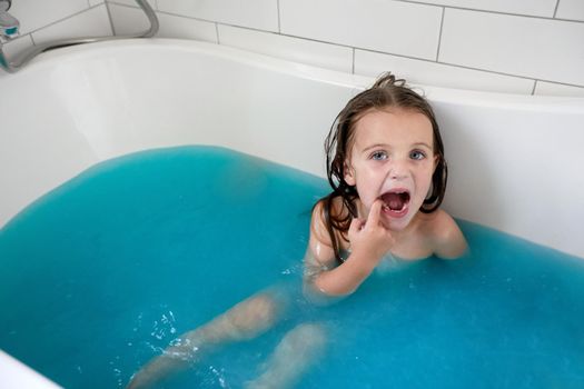 From above of preschooler girl with mouth opened showing teeth while sitting in bathtub with colorful blue water