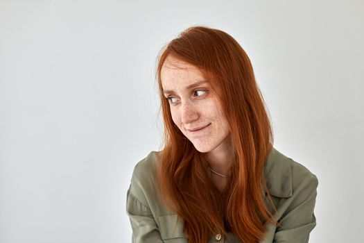 Timid female with long ginger hair and freckles smiling and looking away against white background