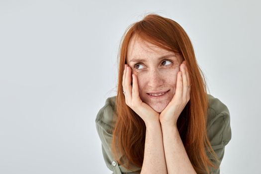 Positive female with ginger hair keeping hands on cheeks and looking away against white background