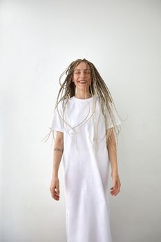 Happy young female with braids wearing casual maxi white dress standing against light wall in studio