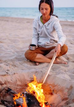 Beach summer vacations, young woman relaxing near a campfire in front of sea.
