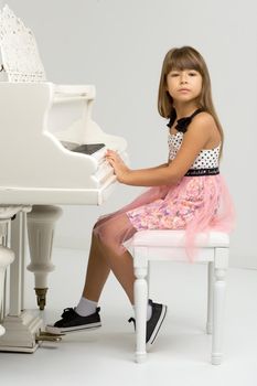 Young girl posing at white grand piano. Adorable girl sitting at at musical instrument. School of arts, music lesson concept. Portrait of preteen child in nice dress looking at camera