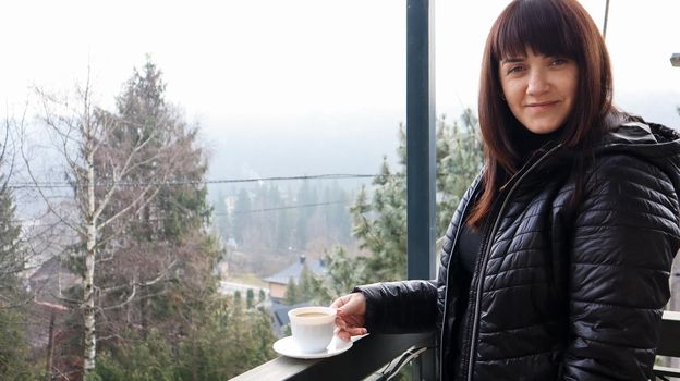 Beautiful happy woman with a white cup of coffee or tea enjoying the beautiful view of the mountain landscape in the morning fog from the balcony of her hotel room. Autumn and winter travel concept.