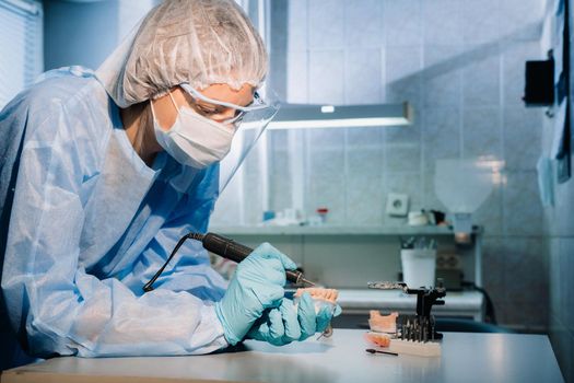 A masked and gloved dental technician works on a prosthetic tooth in his lab.