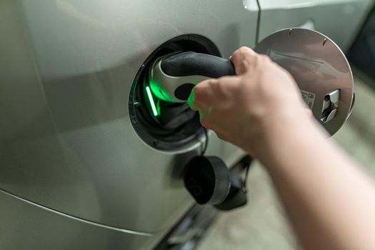 Detail view at a mans hand with is plugin the electric charging wire and plug into his e-automobiles fuel cap. Green light for the active charhing process