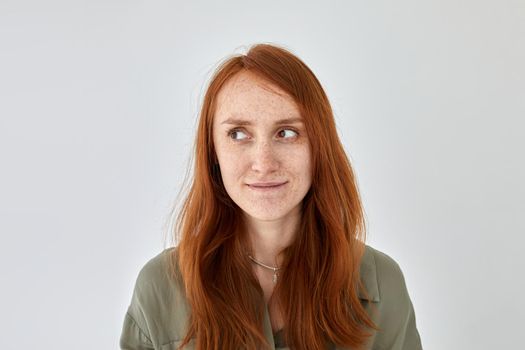 Portrait of glad female with ginger hair and freckles standing on white background and looking away