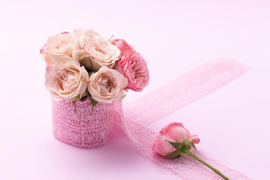 A bouquet of beautiful roses stands in a small bucket on a lace ribbon on a pink background with space for text