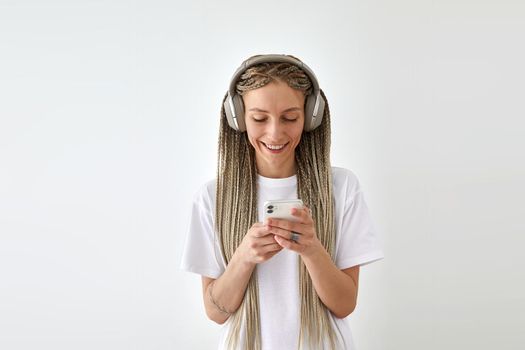 Positive female with long braids wearing white t shirt standing against white background and enjoying songs in headphones while using mobile phone