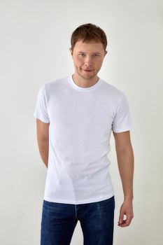 Positive young male in jeans with hand behind and white t shirt standing against white background and looking at camera