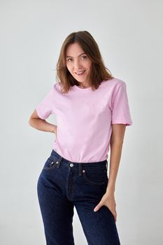 Positive female model wearing pink t shirt and jeans standing with hand on waist against white background and looking at camera