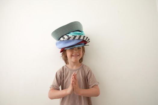 Smiling boy wearing t shirt and various headwear standing against white background and looking at camera. Child's palms are folded at the chest in gratitude