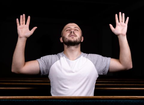 A Christian man in white shirt is sitting with his hands up and praying with humble heart in the church.