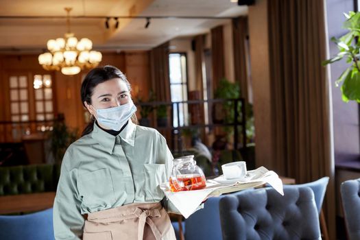 Smiling waitress in mask and apron standing with teapot on tray in cafe and looking at camera