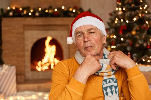 Portrait of mature man wearing orange sweater, scarf and santa claus hat posing in living room at home near christmas tree and fireplace, looking at camera, celebrating new year alone.