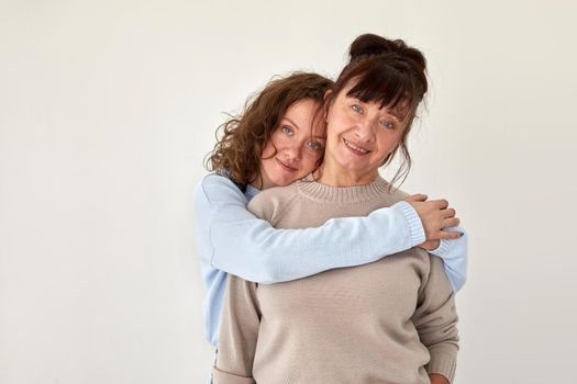 Young woman hugs happy mature smiling lady wearing casual clothing stand together in apartment room with beige wall closeup