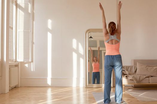 Back view of fit female athlete in sportswear standing in front of mirror at home and stretching raised arms while warming up before workout