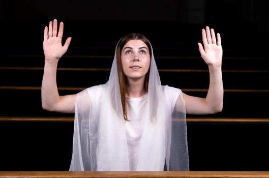 A young modest girl with a handkerchief on her head is sitting in church and praying. The concept of religion, prayer, worship.