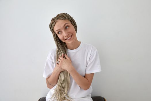 Positive female wearing white t shirt sitting near wall and touching braids while looking away