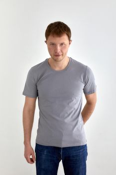 Positive young male in jeans hand behind and gray t shirt standing against white background and looking at camera