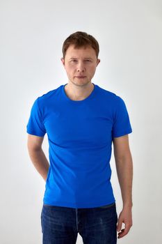 Young male in jeans with hand behind and blue t shirt standing against white background and looking at camera
