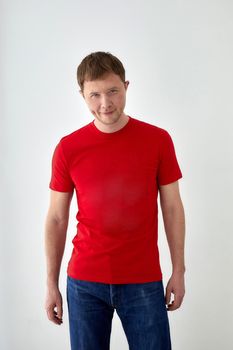 Positive young male model in casual red t shirt and jeans looking at camera while standing against white background