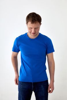 Young male in jeans with hand behind and blue t shirt standing against white background and looking down