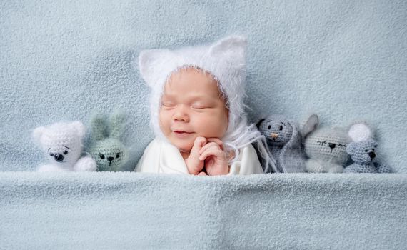 Adorable little infant child in cute bonnet with ears sleeping on the bed with small toys placed near him. Top view