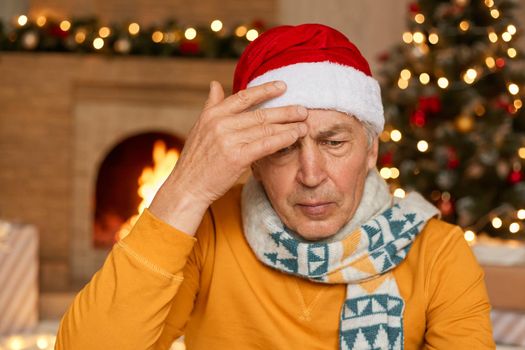 Mature man posing indoor against fireplace and decorated fir tree, keeping hand on forehead, having headache and high temperature, senior male wearing santa hat, scarf and sweater.