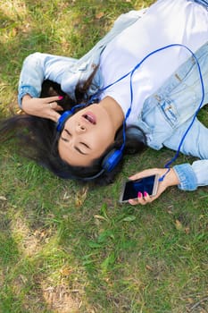 Asian woman enjoying the music while lyuing on the grass in city park - view from the top. She having rest from the office work. Vertical photo.