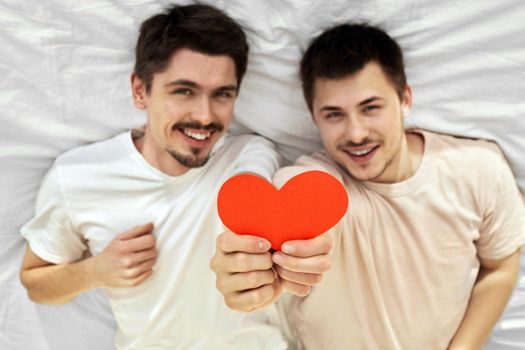 Happy couple of young gay men hide hold red paper heart lying on back on valentine's day in blur