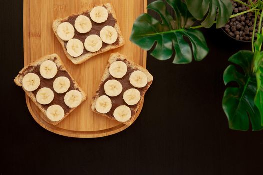 Three banana white bread toasts spread with chocolate butter that lie on a chopping board with a sprig of leaves on a dark background. top view with area for text