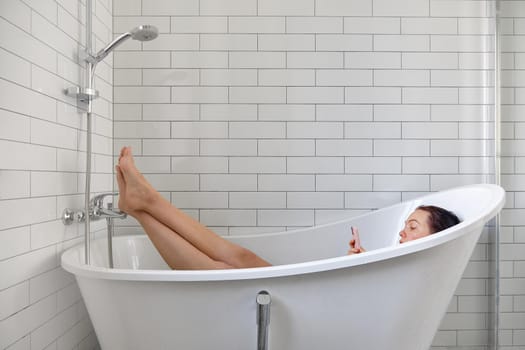 Young female browsing internet on mobile phone while taking bath and relaxing in bathtub