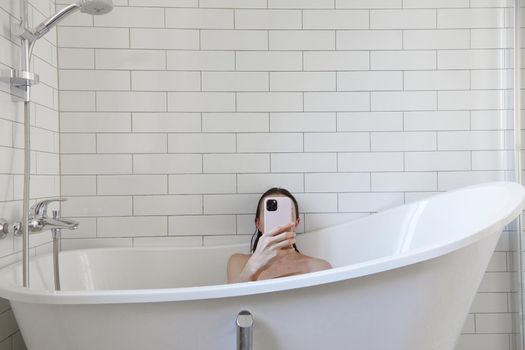 Unrecognizable female covering face with mobile phone while taking bath in white bathtub and surfing internet during daily hygiene routine at home