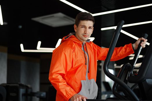 Portrait of a young man in orange windbreaker workout on a fitness machine at a gym.