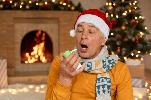 Mature man being ill on Christmas eve, using spray for throat, keeps mouth opened, wearing white jumper, scarf and santa hat, posing in festive living room.