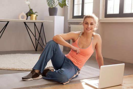 Smiling athletic female in sportswear sitting on mat with laptop during online workout at home and looking at camera