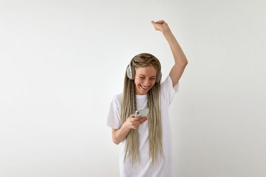 Joyful young female in white shirt listening to music in headphones and dancing with arm raised while using smartphone on white background in studio