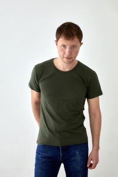 Handsome young male in jeans with hand behind and dark green t shirt standing against white background and looking at camera