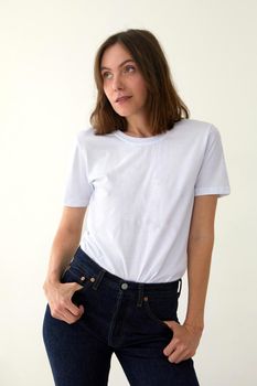 Positive female model wearing white t shirt and jeans standing with hands on waist against white background and looking away