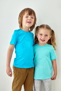 Cheerful toddler girl and preschooler boy in bright blue t shirt hugging and looking at camera while standing against white wall