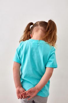 Back view of unrecognizable upset little girl with ponytails dressed in blue t shirt standing against white wall