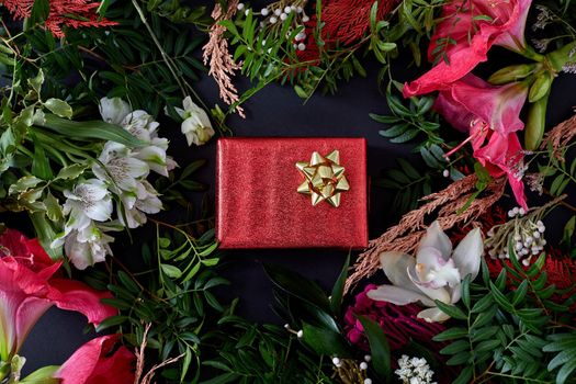 Red box with a gift surrounded by flowers on a dark background flat lay