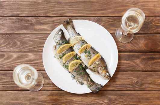 Fish dish with lemons and greenery on a white plate on wooden table with glasses of wine for couple, top view.