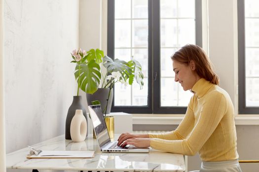 Business woman in yellow turtleneck using laptop computer and sitting by the table in home office