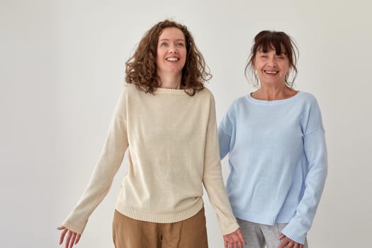 Cheerful middle aged mother and adult daughter standing on gray background in studio and smiling