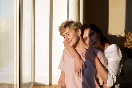 Young woman hugging girlfriend with closed eyes while resting under bright sunlight near window at home