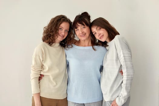Cheerful adult mother embracing charming young daughters while standing on white background in studio and looking at camera