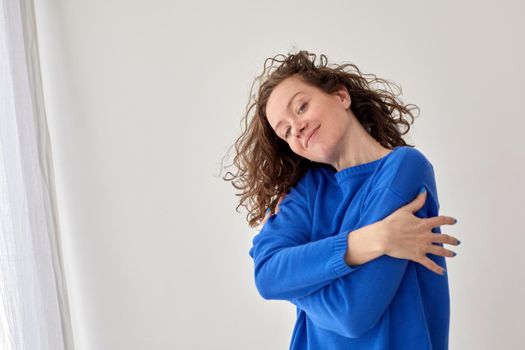 Self hug of happy confident young woman against white wall background. Side view of content female wearing trendy blue sweater standing on gray background studio looking at camera. Girl hugs herself