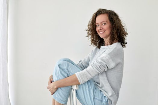 Side view of young female in trendy outfit sitting on wooden chair on white background and looking at camera
